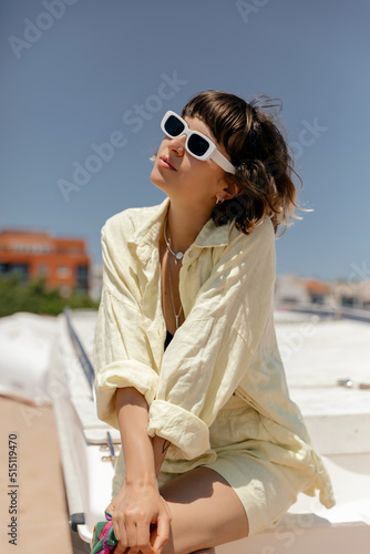 Attractive stylish european short-haired girl in summer outfit in sunglasses sitting on the beach in sunlight and enjoying summer light. Woman is smiling and posing against background of blue sky