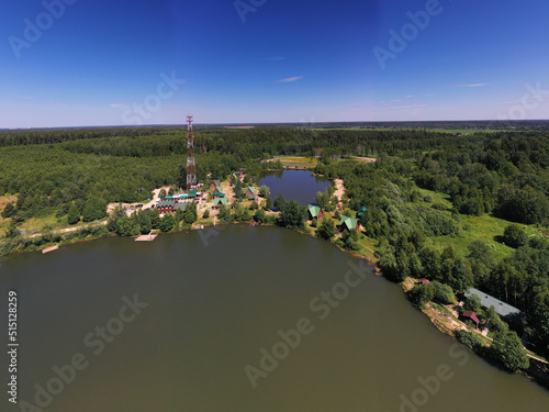 a panoramic view of the green forest and the lake and the glampings standing on the shore on a sunny summer day taken from a drone