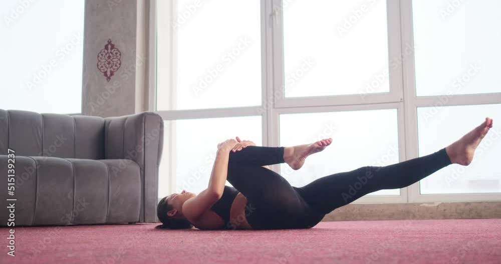 Side view of flexible wearing black top and leggings, lying on red carpet, doing abs, stretching. Pretty, slim female with ponytail raising legs and hands. Concept of new age. Side view of flexible wearing black top and leggings, lying on red carpet, doing abs, stretching. Pretty, slim female with ponytail raising legs and hands. Concept of new age.