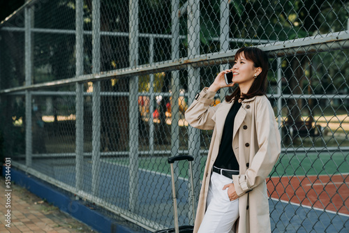 successful asian businesswoman leaning against the fence of basketball court is keeping her hand in the pocket while talking on the phone with luggage. © PR Image Factory
