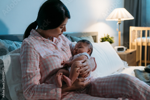 Obraz na płótnie loving asian mother sitting on bed in pajamas is putting her cute baby in her cuddle to sleep at a cozy bedroom during nighttime at home