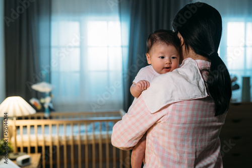 rear view of caring asian mother wearing pajamas with a cloth on shoulder is burping her cute baby daughter in a dark bedroom at night. photo