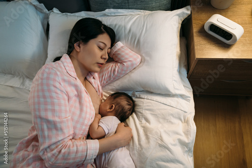 portrait chinese mother wearing pajamas is sleeping on bed while her baby daughter is nestling up to her and suckling milk from breast at bedtime in the bedroom