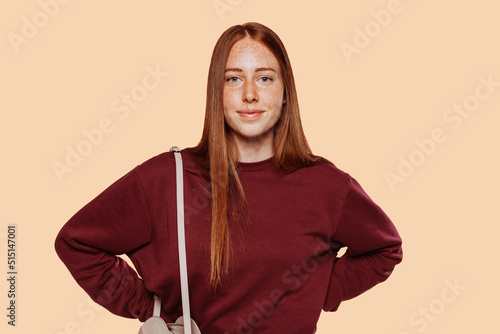 Tablou pe pânză Close up photo of young cheerful red-haired girl she her lady student with freckle and hands on waist, wear red casual sweater and beige bag isolated bright beige background