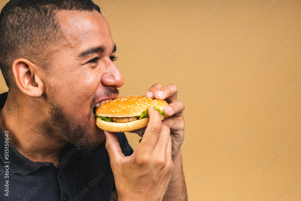 Hungry young african american black man eating hamburger isolated over beige background. Diet or Fast food concept. Hungry young african american black man eating hamburger isolated over beige background. Diet or Fast food concept.