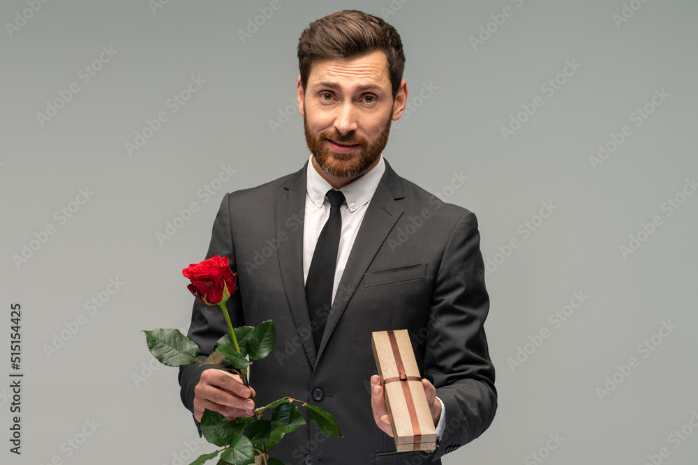Portrait of handsome bearded brunette man in formal suit standing, holding red rose flower and box with present. Indoor studio shot isolated on grey background Portrait of handsome bearded brunette man in formal suit standing, holding red rose flower and box with present. Indoor studio shot isolated on grey background
