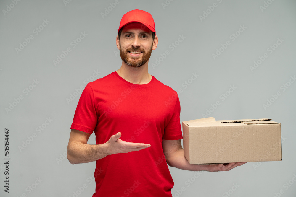 Waist up view portrait of male courier in red uniform handing cardboard parcel to client, carrying parcel with goods ordered online. Indoor studio shot isolated on grey background Waist up view portrait of male courier in red uniform handing cardboard parcel to client, carrying parcel with goods ordered online. Indoor studio shot isolated on grey background