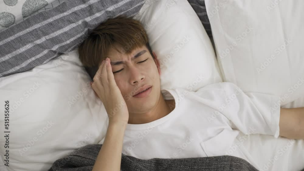 high angle view with asian man lying awake on bed is rubbing his temples with a frown while suffering a bad headache in the morning at home. high angle view with asian man lying awake on bed is rubbing his temples with a frown while suffering a bad headache in the morning at home.