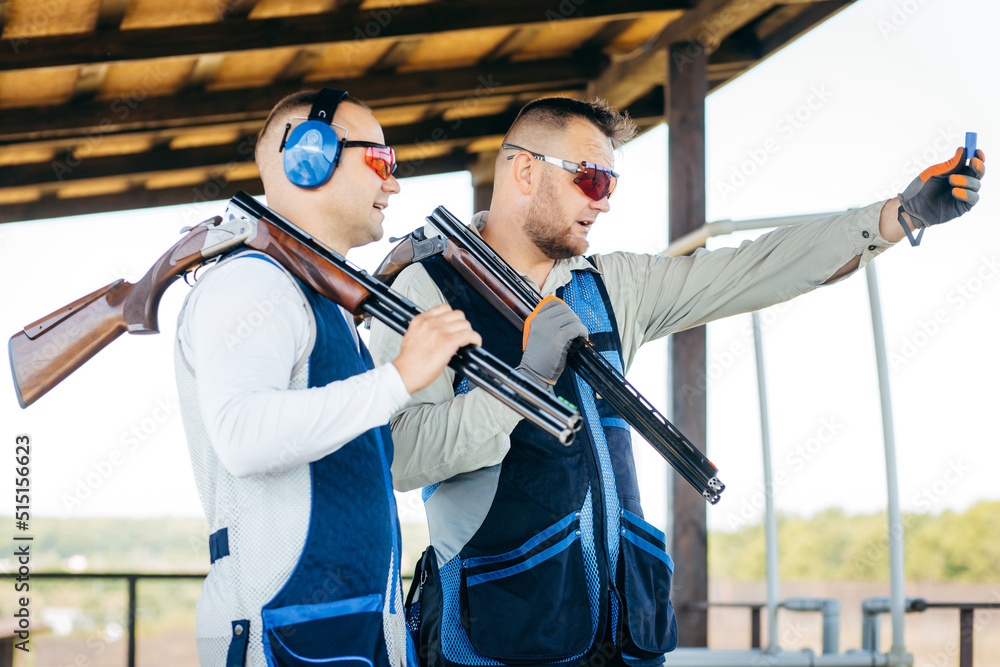 Two adult mans in sunglasses and a rifle vest practicing fire weapon shooting. Two young experienced shooters aiming shotgun in outdoor.
