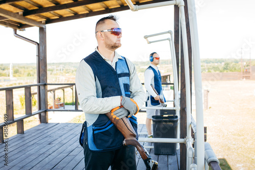 Two adult mans in sunglasses and a rifle vest practicing fire weapon shooting. Two young experienced shooters aiming shotgun in outdoor. © andrew_shots