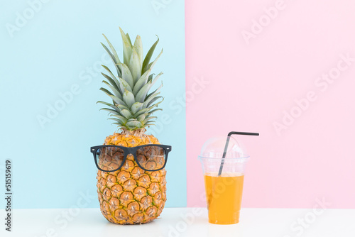 Fresh pineapple with sunglasses near a glass of juice and a cocktail straw on color background