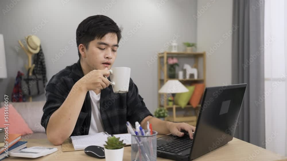 asian male freelance worker nodding head while having a short tea break and continuing working on the project on the computer at home. asian male freelance worker nodding head while having a short tea break and continuing working on the project on the computer at home.