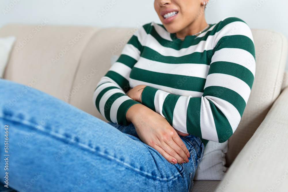 Fototapeta premium African American Woman in painful expression holding hands against belly suffering menstrual period pain, lying sad on home bed, having tummy cramp in female health concept