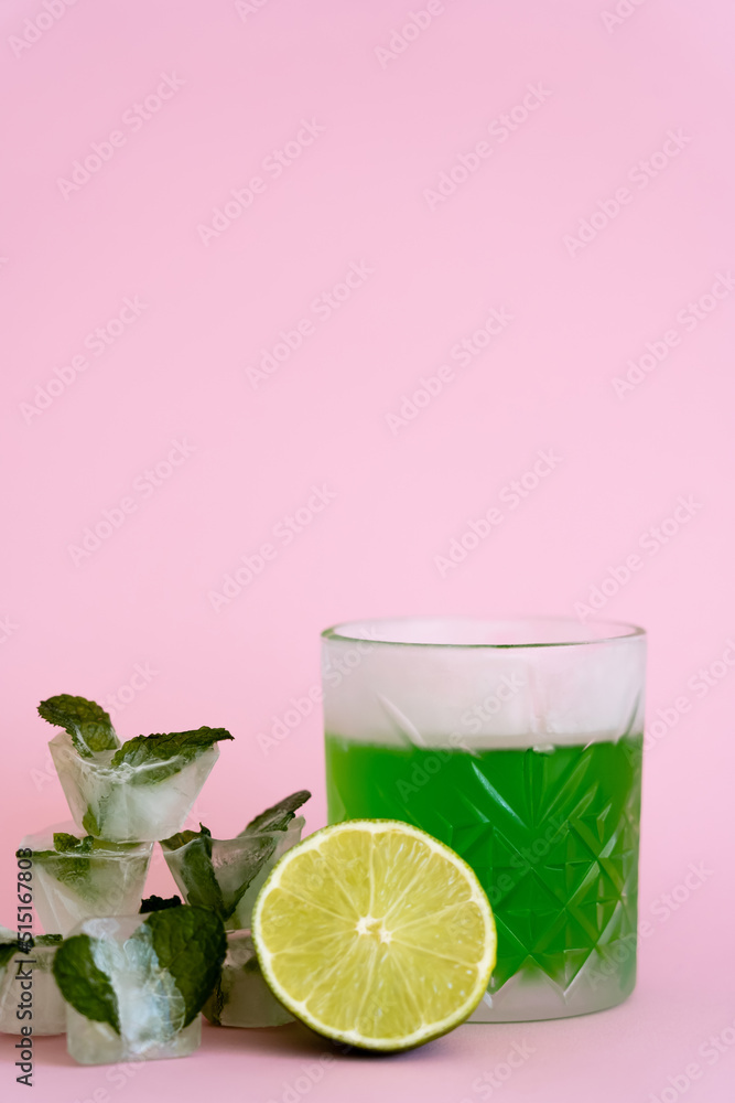 green and fresh lime in faceted glass with alcohol drink near ice cubes with peppermint on pink. green and fresh lime in faceted glass with alcohol drink near ice cubes with peppermint on pink.