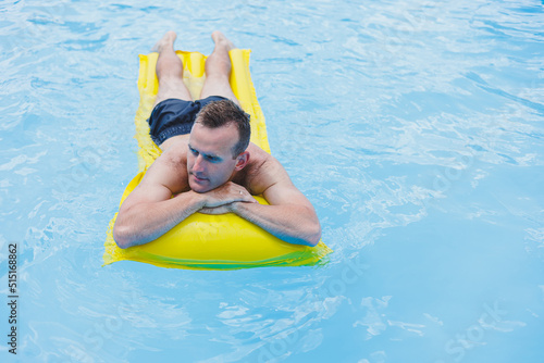 A young man in shorts enjoys the water park floating in an inflatable big ring on a sparkling blue pool smiling at the camera. Summer vacation.