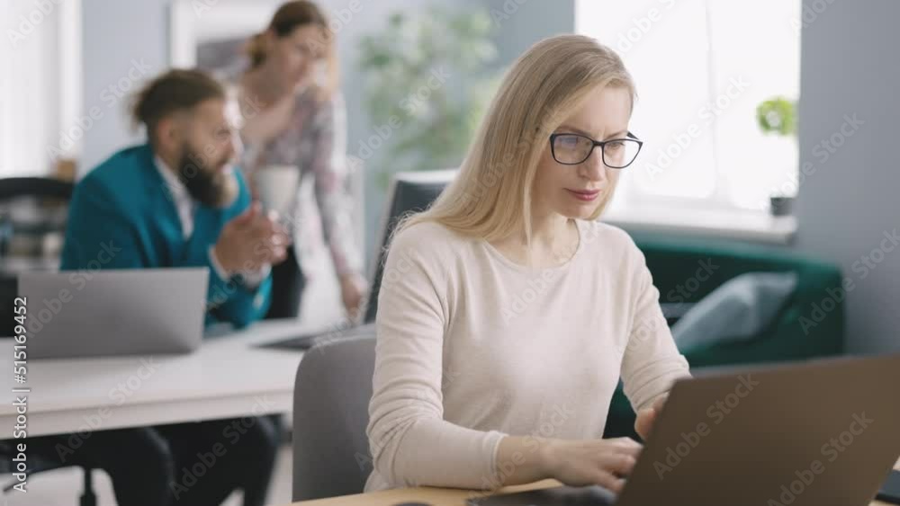 Charming woman using laptop in bright office with colleagues on background. Confident specialist in casual clothes analyzing statistical data in charts. Charming woman using laptop in bright office with colleagues on background. Confident specialist in casual clothes analyzing statistical data in charts.