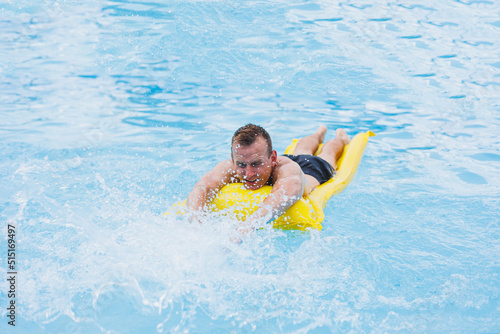 A young man in shorts enjoys the water park floating in an inflatable big ring on a sparkling blue pool smiling at the camera. Summer vacation.
