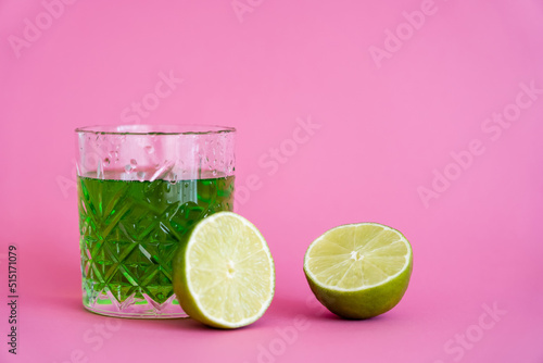 green alcohol drink in faceted glass with water drops near limes on pink.