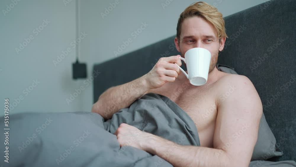 Happy blond athletic man with naked torso drinking coffee while lying on the bed in the morning Happy blond athletic man with naked torso drinking coffee while lying on the bed in the morning