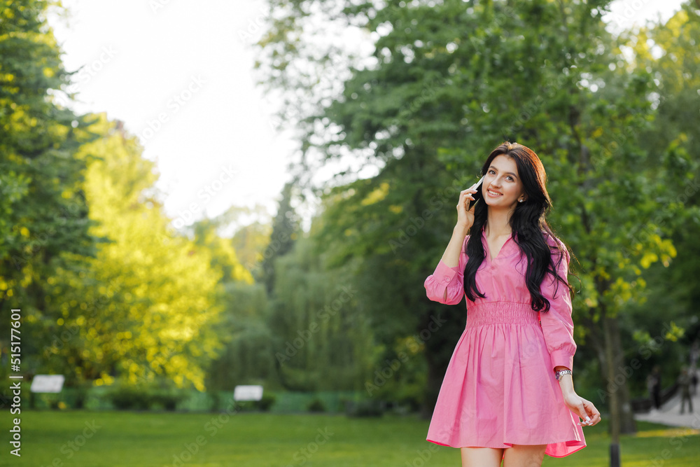 Close up portrait of a young brunette with wavy hair dressed in a pink dress walking in the park talking on a cell phone. Close up portrait of a young brunette with wavy hair dressed in a pink dress walking in the park talking on a cell phone.