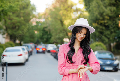 Street style, fashion concept: elegant woman in fashionable pink dress, white hat, posing on city street, looking at watch on hand