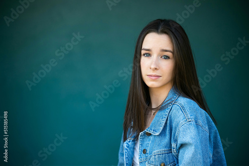 A beautiful teen brunette girl with a serious look leaning against a wall in thought with a teal green turquoise background