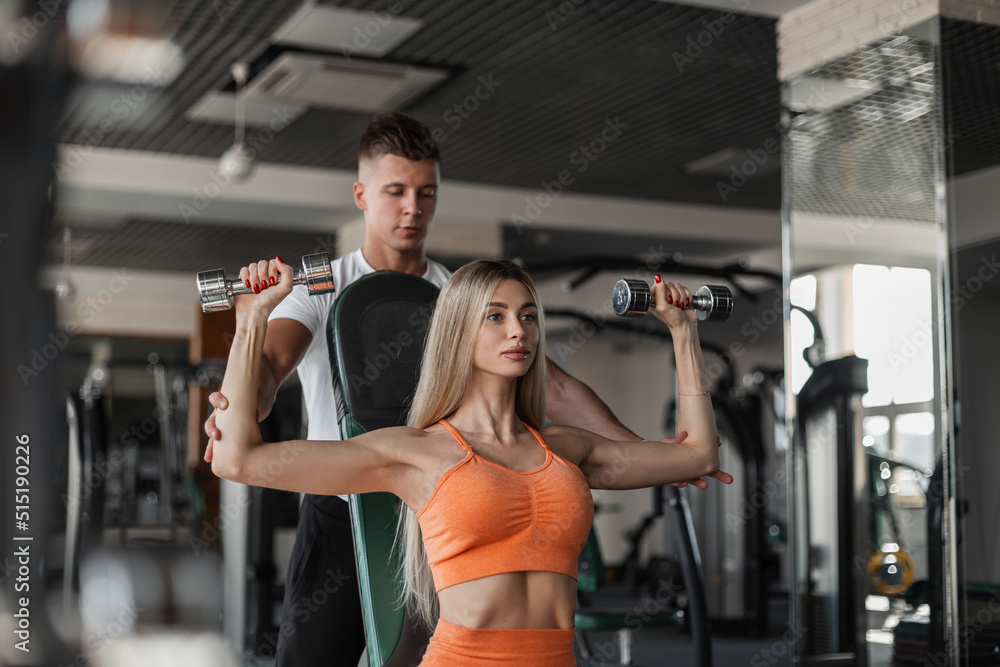 Athletic beautiful young couple working out in the gym with dumbbells. Beautiful blonde woman with fitness trainer works out her body Athletic beautiful young couple working out in the gym with dumbbells. Beautiful blonde woman with fitness trainer works out her body