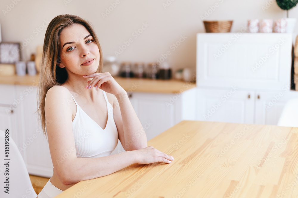 Young woman is sitting at Wooden Table in front of White Cozy Kitchen. Rent Housing with Good Repair Young woman is sitting at Wooden Table in front of White Cozy Kitchen. Rent Housing with Good Repair