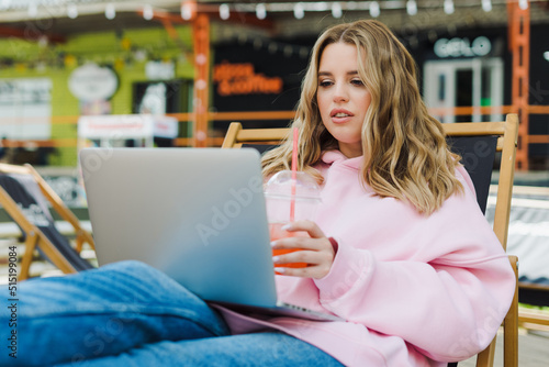 A young freelance woman is working on a laptop and drinking lemonade in a street cafe. A model in a pink hoodie and jeans