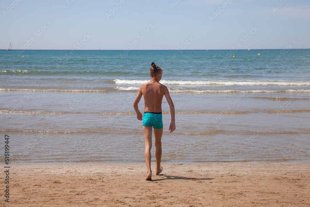 A teenager enters the sea, view from the back. Young boy, view from the back, looks at the Mediterranean sea. Summer holiday vacation travel concept A teenager enters the sea, view from the back. Young boy, view from the back, looks at the Mediterranean sea. Summer holiday vacation travel concept
