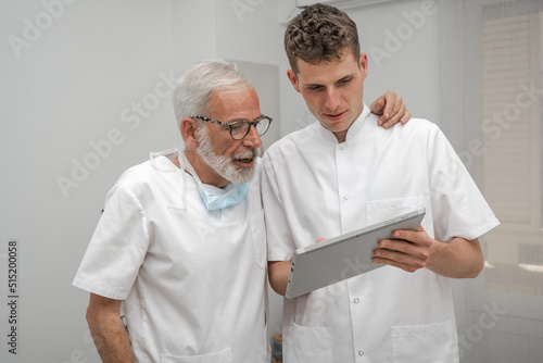 Mature smiling doctor and young intern doctor discussing patient diagnosis, holding digital tablet. Father and son dentist doctors