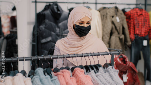 Young arab woman shopper in medical protective mask chooses clothes in clothing store retail sales discount purchase shopping in boutique muslim female customer in hijab looking for new warm sweater
