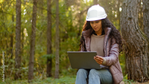 Attractive woman forestry engineer in protective helmet enters data into laptop takes reforestation action young experienced female specialist ecologist technician watching nature reserve checks trees photo
