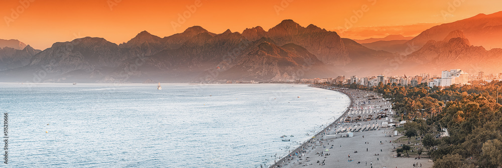 Naklejka premium Sunset panoramic view of scenic and popular Konyaalti beach in Antalya resort town. Majestic mountains with haze in the background. Vacation and holiday in Turkey