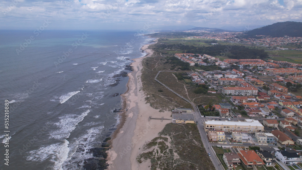 Fototapeta premium Aerial view of the Northern Littoral Natural Park in Esposende, Portugal. Sea, beach boulders, pebble shore, and waves. Sea waves breaking on rocks. Sunny beach with sand dunes and a cloudy sky.