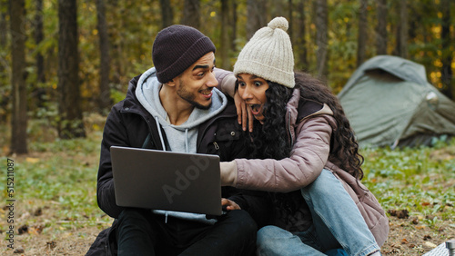 Young happy couple in love tourists sit outdoors in campsite look at laptop screen choose place to travel on electronic map use computer application backpackers communicate laughing enjoy vacation