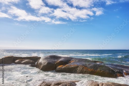 Landscape of large rocks in the ocean with a cloudy blue sky and copy space. Sea waves splashing against boulders on popular Camps Bay beach in Cape Town, South Africa. A beautiful summer destination