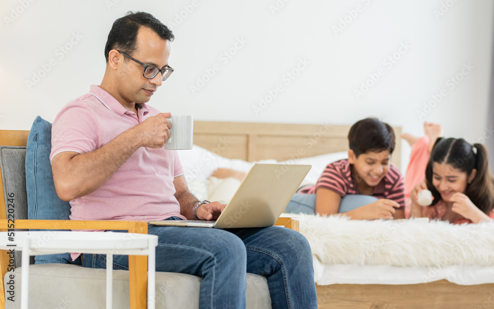 Indian father working at home or apartment, drinking coffee in morning, reading news, using laptop with blur background of children playing on bed. Family, Lifestyle, Business, Technology Concept. Indian father working at home or apartment, drinking coffee in morning, reading news, using laptop with blur background of children playing on bed. Family, Lifestyle, Business, Technology Concept.