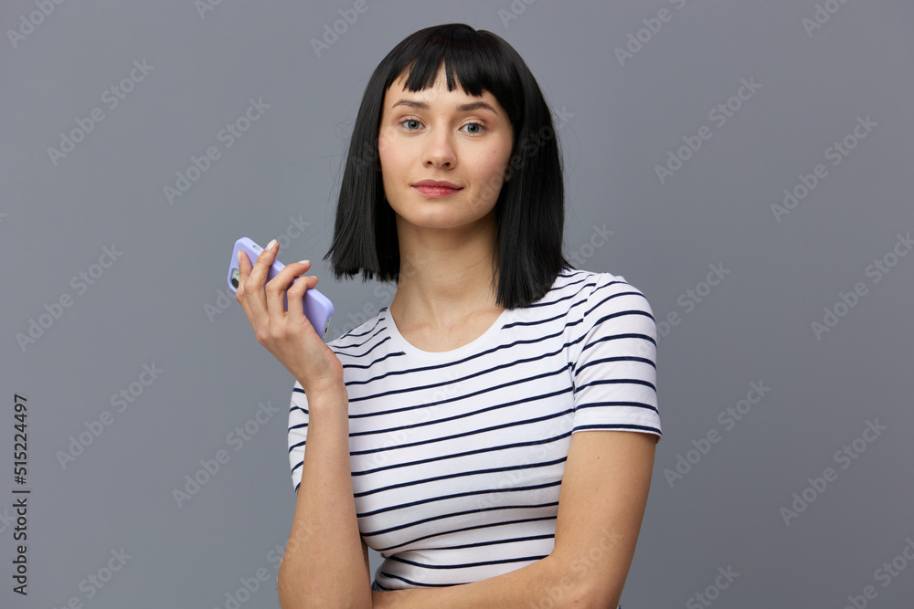 an attractive, beautiful woman with short black hair stands on a gray background in a striped cotton T-shirt and holds a smartphone in her hand looking at the camera. an attractive, beautiful woman with short black hair stands on a gray background in a striped cotton T-shirt and holds a smartphone in her hand looking at the camera.