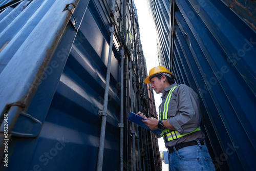 Asian male worker standing between containers, lookinng and Check sets to write reports, to workers and Cargo container concept.