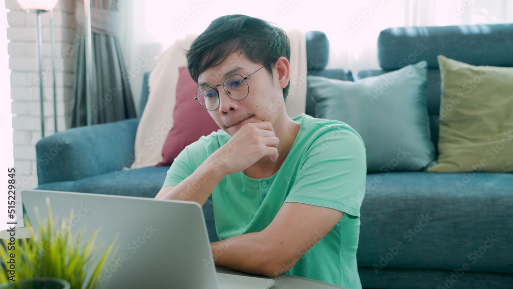 Naklejka premium Close-up of a young Asian man in a green shirt, sitting at a laptop screen, with a serious look on his face and a thoughtful expression.