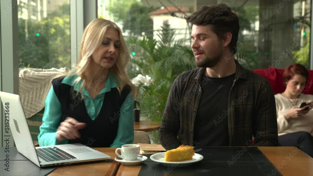 Older woman explaining work to younger colleague at coffee shop showing laptop and taking notes Older woman explaining work to younger colleague at coffee shop showing laptop and taking notes