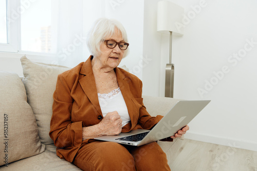a serious, experienced, elderly woman is sitting working from home on the couch with a laptop on her lap and menacingly looks at the monitor with her hand in a fist © Tatiana