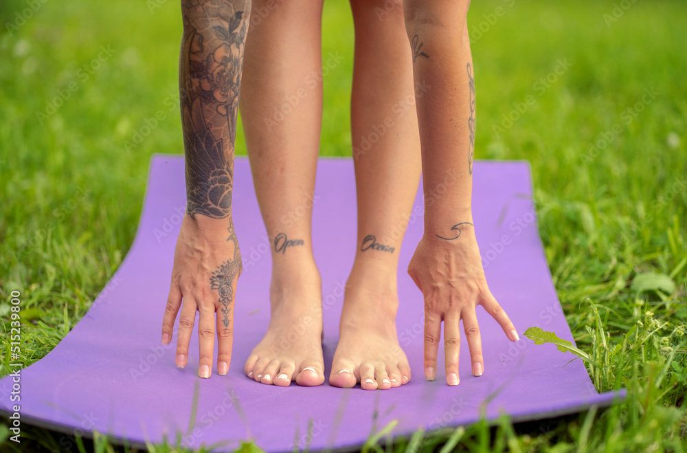 The girl stands on a yoga mat in the park and stretches her hands to her feet