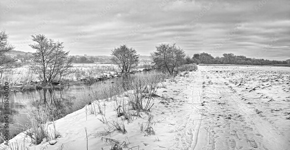 Obraz premium A black and white view of a mountain lake in winter, covered with snow and ice. The beautiful cloudy weather around the pond covered with snowfall. A dense forest and cloudy sky on a glacial valley.