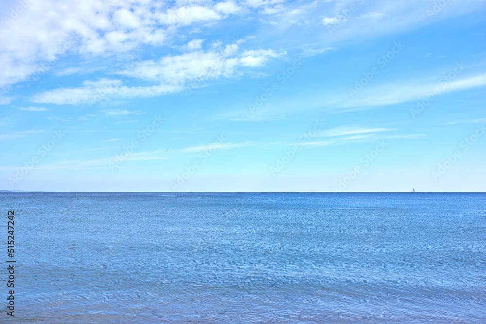 Copy space at sea with a cloudy blue sky background. Calm ocean waves at an empty beach with a sailboat cruising in the horizon. Scenic and picturesque landscape view for a peaceful summer holiday