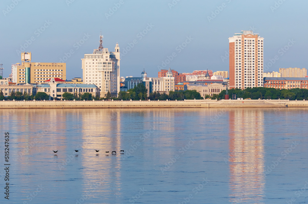 Naklejka premium View of China from Russia across the Amur River on a summer morning. A flock of cormorant birds are flying over calm water. City buildings of different styles and purposes. Clear sky.