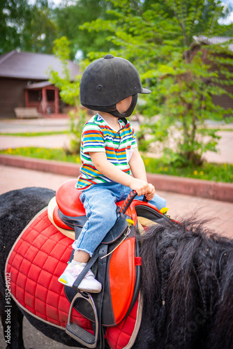 Beautiful little girl two years old riding pony horse in big safety jockey helmet posing outdoors on countryside 