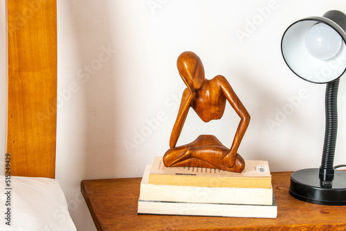 wooden night table, with black lamp, books and a small wooden human figure sitting on it. 