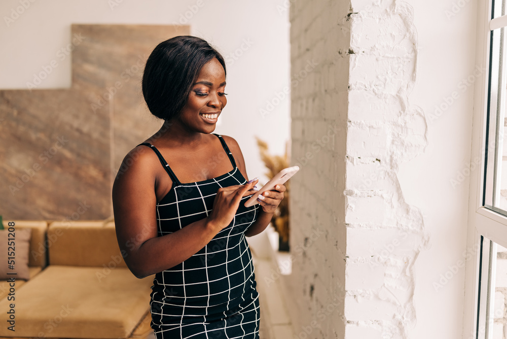 Obraz premium Cheerful young woman using smartphone. Smiling woman using app on cellphone at home. Beautiful girl relaxing while chatting on mobile phone.